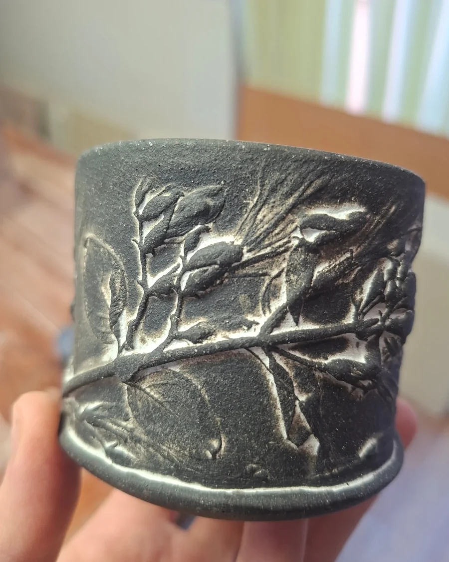 Dark clay beaker held in one hand, banded with a raised botanical relief
